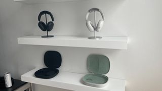 Two pairs of Sonos Ace headphones (one white, one black) on stands on a shelf with their open travel cases on the shelf below.