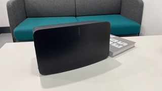 A black Sonos Five wireless speaker on a white table in front of a hardback book and a green and grey sofa.