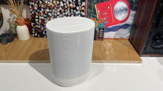 A white Sonos Move 2 on a white surface in front of a wooden shelf with an incense burner and artworks.