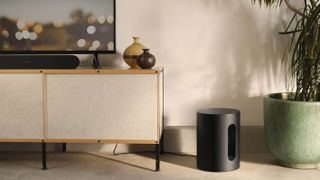 A black Sonos Sub Mini on the floor next to a white TV cabinet bearing a TV. Next to the Sub Mini is a green plant pot with a tall plant growing out of it.