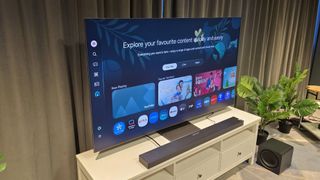 Samsung QN990F 8K TV sitting on a white media unit with a soundbar in front and a plant on the right hand side.